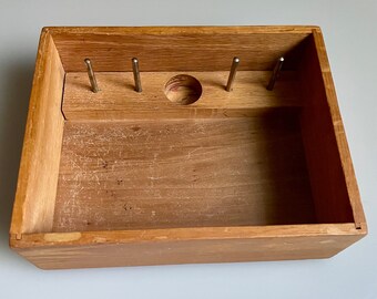 May include: A wooden box with a small hole in the center and four metal pegs on the top. The box is empty.