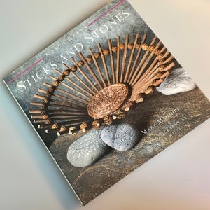 May include: Book cover with the title "Sticks and Stones: New Crafts" by Mary Maguire. The cover features a woven basket made of sticks and stones, with a brown background.