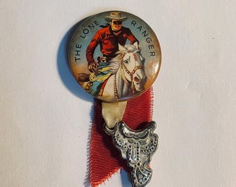 May include: A vintage pin featuring an illustration of the Lone Ranger riding a white horse, with the text "THE LONE RANGER" around the edge. The pin is attached to a red ribbon and a silver-colored saddle charm.
