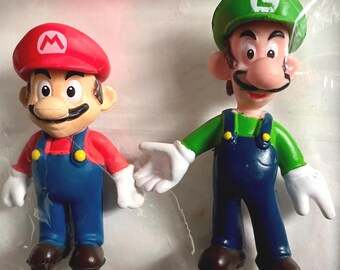 May include: Two plastic figurines of Mario and Luigi from the Super Mario Bros. video game series. Mario is wearing a red hat with an 'M' on it, a red shirt, blue overalls, and brown shoes. Luigi is wearing a green hat with an 'L' on it, a green shirt, blue overalls, and brown shoes.