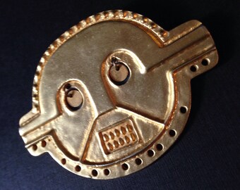 May include: A gold-colored, stylized metal artifact with a circular shape and geometric design. The piece features two circular cutouts and a rectangular section with small, evenly spaced rectangles. The edges are adorned with small, evenly spaced holes.