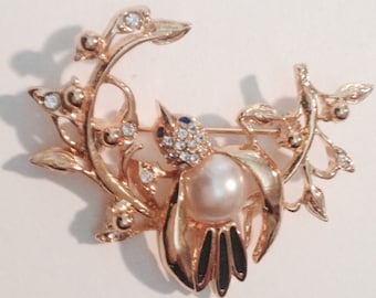 May include: A gold-tone brooch featuring a bird perched on a branch with a pearl accent. The bird is adorned with clear rhinestones and blue enamel accents.