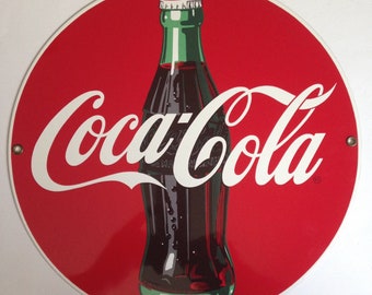 May include: Round red Coca-Cola sign with a white border. The sign features a green and black illustration of a Coca-Cola bottle in the center, with the Coca-Cola logo in white script across the front.