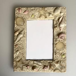 May include: A rectangular picture frame with a textured, sandy-colored surface decorated with various seashells and starfish. The frame features a central vertical rectangular opening with a white insert, surrounded by shells of different shapes.