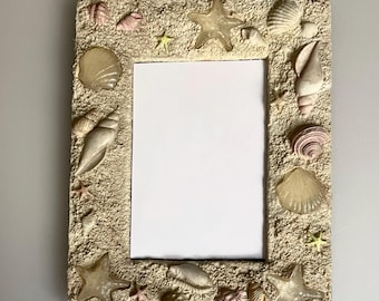 May include: A rectangular picture frame with a textured, sandy-colored surface decorated with various seashells and starfish. The frame features a central vertical rectangular opening with a white insert, surrounded by shells of different shapes.