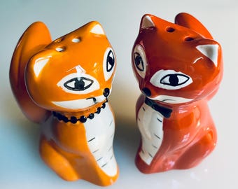 May include: A pair of ceramic fox-shaped salt and pepper shakers. One is orange with a black beaded necklace, the other is red with a black bow tie. Both have black and white facial details and holes on top for dispensing.