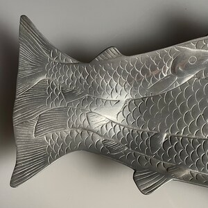 May include: A silver fish-shaped serving dish with a detailed design of overlapping fish scales. The dish is made of metal and has a smooth, polished finish.
