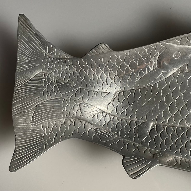 May include: A silver fish-shaped serving dish with a detailed design of overlapping fish scales. The dish is made of metal and has a smooth, polished finish.
