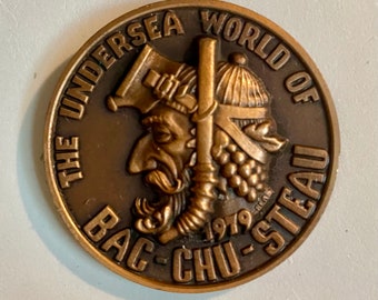 May include: A bronze coin with the text "The Undersea World of Bac-Chu-Steau 1979" around the edge. The center of the coin features two cartoon faces with a large axe and a smaller axe.