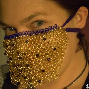May include: A gold chainmail mask with purple trim covers the lower half of a person's face. The mask is made of small, interlocking metal rings and has a decorative pattern.