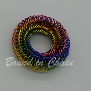 Puede incluir: Un anillo de malla de cadena de color arcoíris hecho de anillos de metal. El anillo tiene un diámetro de aproximadamente 5 cm y está hecho de una variedad de colores, incluyendo rojo, naranja, amarillo, verde, azul y morado.