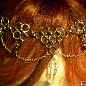 May include: A silver chainmail crown with a crystal and black beads hanging from it. The crown is worn on a person with red hair.
