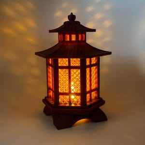 May include: A dark brown, illuminated Japanese pagoda lantern. The lantern has a multi-tiered roof and a geometric lattice design on the sides. The interior glows with warm orange light, creating a cozy ambiance. The lantern sits on a hexagonal base.