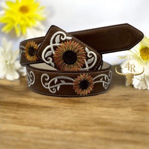 Puede incluir: Un cinturón de cuero marrón con girasoles bordados y volutas blancas. El cinturón presenta un extremo de hebilla rectangular y una punta. El diseño incluye el texto "MANOLO BOOTS 4R".