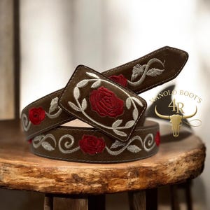 Puede incluir: Un cinturón de cuero marrón con bordados de rosas rojas y detalles blancos. La hebilla rectangular presenta una gran rosa bordada y un extremo decorativo. La marca "MANOLO BOOTS 4R" es visible.