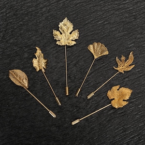 May include: Six gold-colored lapel pins in various leaf designs. Each pin features a detailed leaf shape attached to a long, slender pin with a secure clasp. The pins are arranged on a dark, textured surface.