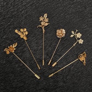 May include: Six gold-coloured lapel pins with floral designs. The pins feature various flower and leaf motifs, with long, slender stems and decorative tops. The pins are displayed on a dark, textured surface.