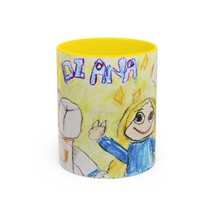 May include: A white ceramic mug with a yellow interior. The exterior features a child's drawing of a person with blonde hair and a blue dress, and the name "DIANA" in blue letters. The background is yellow with orange diamond shapes.