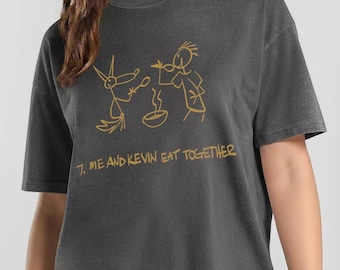 Puede incluir: Una camiseta de manga corta en color gris carbón con un dibujo lineal dorado de un zorro y una persona comiendo de un cuenco. Debajo de la ilustración, el texto dice: 7. ME AND KEVIN EAT TOGETHER. La prenda tiene cuello redondo y corte holgado.