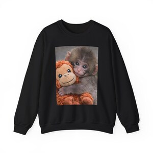 Può includere: Felpa nera con una grafica di una scimmia che abbraccia un peluche di un orango. La scimmia ha la pelliccia grigia e il peluche è arancione. L'immagine è centrata sulla parte anteriore della felpa.