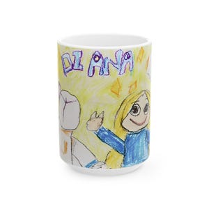 May include: White ceramic mug featuring a child's drawing. The artwork includes the name "DIANA" in purple letters, a smiling girl in blue, and a figure in white. The background is yellow with orange diamond shapes.