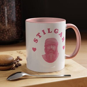 Può includere: Una tazza in ceramica bianca con interno e manico rosa. La parte anteriore presenta una grafica rosa di una persona che indossa un copricapo e una maschera, con il testo STILGAR arcuato sopra e due piccoli cuori rosa ai lati.