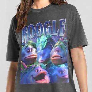 Può includere: T-shirt grigio scuro con una grafica di creature blu simili a cartoni animati con labbra grandi e occhi espressivi. La parola "BOOGLE" è stampata sopra le creature in un carattere blu stilizzato con uno sfondo di fulmini. La maglietta è uno stile casual e unisex.