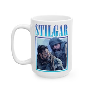 Puede incluir: Una taza de cerámica blanca con un estampado gráfico en tonos azules de dos personajes de la serie de películas Dune. El nombre STILGAR está impreso en letras azules grandes sobre la imagen, que muestra a dos hombres barbudos.