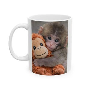 May include: White ceramic mug featuring a smiling baby monkey hugging a stuffed toy monkey. The plush toy is brown with a smiling face. The mug is a standard size and has a white handle.