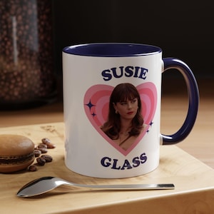 May include: A white ceramic mug with a dark blue interior and handle. The front features a pink heart graphic with a portrait of a woman, surrounded by small star icons. The text SUSIE is printed above the heart and GLASS is printed below.