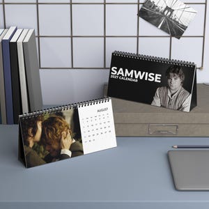 May include: A spiral-bound desk calendar titled Samwise 2027 Calendar featuring a black and white portrait of a man on the cover and a color photograph of two people embracing on the August page with a clear monthly grid.