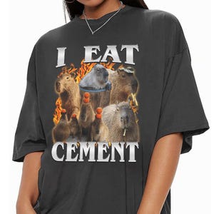 May include: A charcoal grey short-sleeve t-shirt featuring a graphic print of multiple capybaras, some with small orange fruits balanced on their heads, set against a background of orange flames with the text I EAT CEMENT in silver lettering.