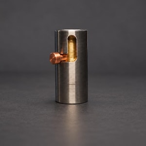 May include: A cylindrical metal object with a brushed silver finish featuring a vertical cutout slot. A small copper-colored pin is positioned within the slot, allowing for movement along the length of the opening in the side of the cylinder.