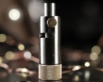 May include: A stainless steel bottle stopper with a textured, knurled base and a small, round, brass-colored accent. The stopper has a cylindrical design with a small lever and a top section. The background is dark with bokeh lights.