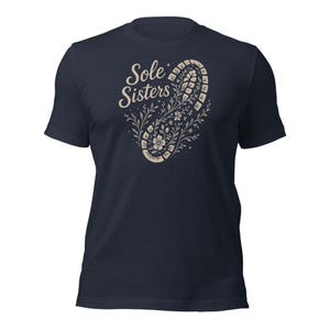 May include: Navy blue t-shirt with the text "Sole Sisters" in a light tan script. A floral design in the shape of a footprint is below the text, with flowers and leaves in a similar tan color. The shirt is made of a soft material.