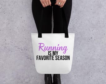 Puede incluir: Bolso tote blanco con asas negras con el texto "Running is my favorite season" en morado y negro. El bolso lo sostiene una persona que lleva pantalones y botas negras.