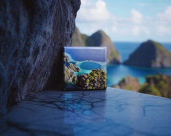 May include: A small square canvas print of a tropical island with a turquoise lagoon. The print is sitting on a gray stone surface with a blurred background of a tropical island.