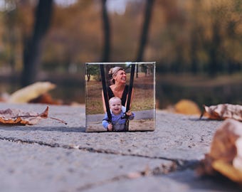 May include: A small, square canvas print featuring a smiling woman and baby on a swing set. The print is set on a gray stone surface with fallen leaves in the foreground, and a blurred park background.