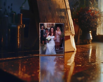 Pode incluir: Uma fotografia emoldurada de um grupo de casamento. A noiva está usando um vestido branco e segurando um buquê de flores brancas. As outras pessoas na foto estão usando roupas formais.