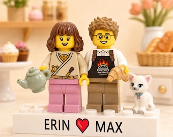 May include: Two custom LEGO minifigures standing on a white rectangular brick base with the text ERIN, a red heart symbol, and MAX printed on the front. The figures include a brown-haired person holding a teapot and a person with glasses wearing a black apron with a burger graphic holding a croissant, accompanied by a small white cat figure.