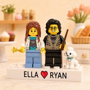 Puede incluir: Dos minifiguras LEGO personalizadas sobre una base rectangular blanca con el texto ELLA, un símbolo de corazón rojo y RYAN impreso en el frente. La figura de la izquierda tiene cabello castaño, chaqueta blanca y varita dorada. La figura de la derecha tiene cabello negro, gafas, camisa negra estampada y varita marrón, acompañada de una pequeña figura de caniche blanco.