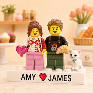 Könnte beinhalten: Zwei individuelle LEGO-Minifiguren auf einem weißen, rechteckigen Sockel mit dem aufgedruckten Text AMY, einem roten Herzsymbol und JAMES. Die linke Figur hat braunes Haar, ein weiß-rotes Oberteil und rosa Beine und hält ein rosa Herz. Die rechte Figur hat braunes Haar, ein schwarzes T-Shirt mit Burger-Grafik und grüne Beine und hält ein Croissant. Eine kleine weiße Katzenfigur sitzt daneben.