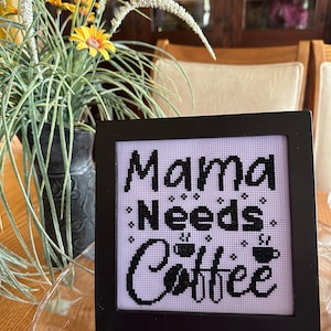 May include: A framed cross-stitch with the text "Mama Needs Coffee" in black thread on a light purple background. The frame is black and square. The image is on a glass plate with a vase of yellow flowers in the background.