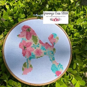 May include: A cross-stitch hoop art featuring a poodle silhouette filled with pink, green, and teal floral patterns. The wooden hoop is set against a backdrop of green foliage. The text "Grammy's Cuss Stitch" is visible.