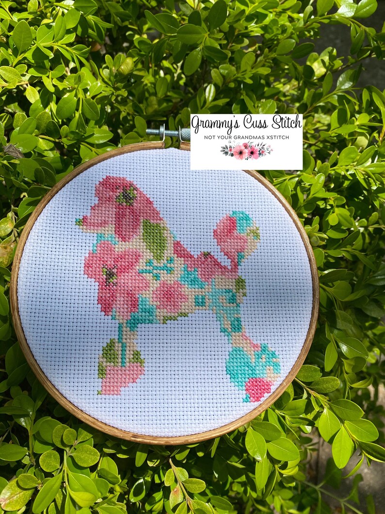 May include: A cross-stitch hoop art featuring a poodle silhouette filled with pink, green, and teal floral patterns. The wooden hoop is set against a backdrop of green foliage. The text "Grammy's Cuss Stitch" is visible.