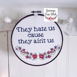 K&ouml;nnte beinhalten: Ein Stickrahmen mit dem Spruch "They hate us cause they ain't us" in dunkelblauer Stickerei. Der untere Teil ist mit einem Blumengirlande in Rot-, Rosa-, Gelb- und Lilat&ouml;nen verziert. Auf wei&szlig;em Hintergrund.