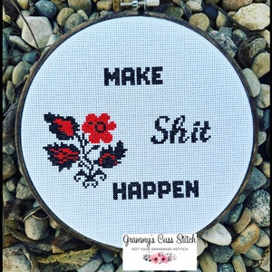 K&ouml;nnte beinhalten: Ein Kreuzstich-Stickrahmen mit den Worten "MAKE Shit HAPPEN" in schwarzen Blockbuchstaben. Auf der linken Seite befindet sich ein rot-schwarzes Blumendesign. Der Rahmen ist vor einem Hintergrund aus Steinen platziert.