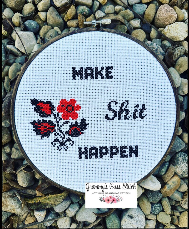 K&ouml;nnte beinhalten: Ein Kreuzstich-Stickrahmen mit den Worten "MAKE Shit HAPPEN" in schwarzen Blockbuchstaben. Auf der linken Seite befindet sich ein rot-schwarzes Blumendesign. Der Rahmen ist vor einem Hintergrund aus Steinen platziert.