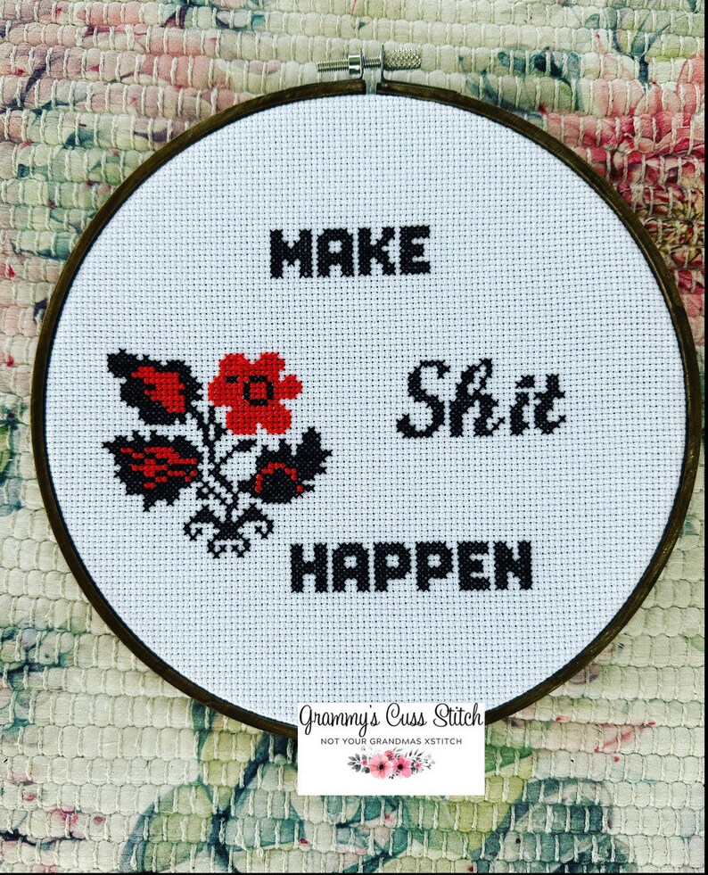 K&ouml;nnte beinhalten: Kreuzstich-Stickerei in einem Holzreifen. Das Design zeigt die Worte "MAKE Shit HAPPEN" in schwarzen Blockbuchstaben, mit einem roten und schwarzen Blumenmotiv. Der Text "Grammy's Cuss Stitch" befindet sich unten.