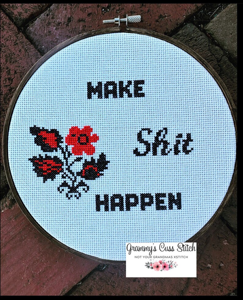 K&ouml;nnte beinhalten: Kreuzstich-Stickerei in einem Holzrahmen mit den Worten "MAKE SHIT HAPPEN" in schwarzen Blockbuchstaben. Ein Blumenmuster in Rot und Schwarz befindet sich auf der linken Seite. Der hellblaue Stoff ist in einem h&ouml;lzernen Stickrahmen befestigt.